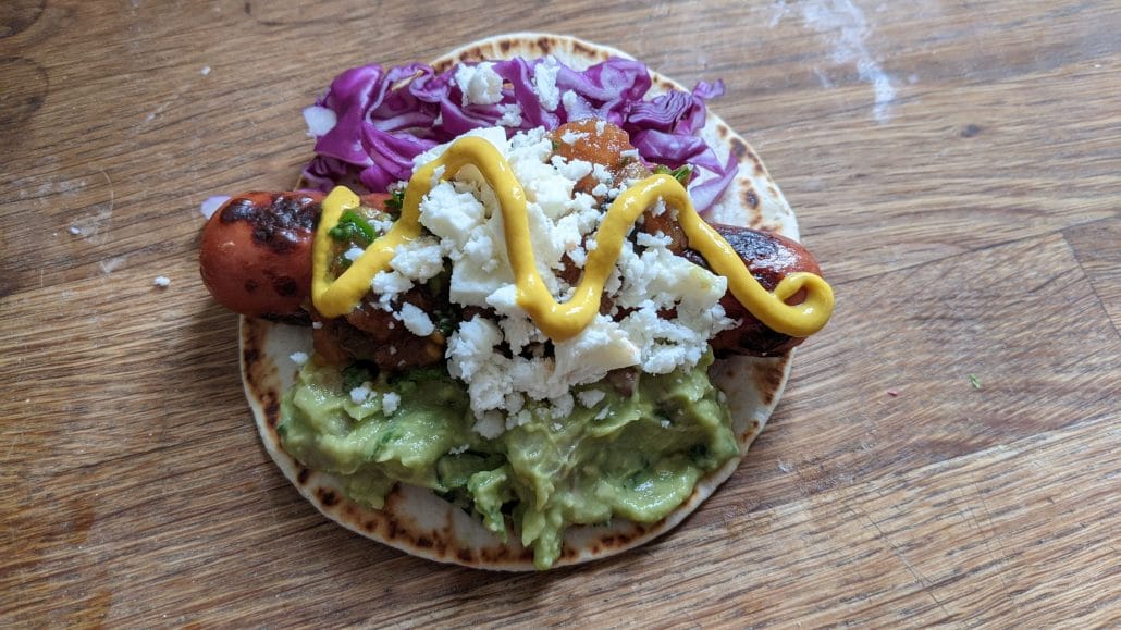 Finished mixtas, a hot dog with guacamole, cabbage, hot sauce, cheese, and mustard on a soft tortilla.